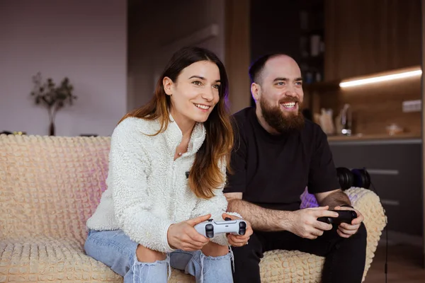 stock image A young couple is sitting on the sofa and playing on a video game console. They look at each other with smiles on their faces and have fun together. Everyone holds their controller in hand and concentrates on the game, trying to defeat the opponents.