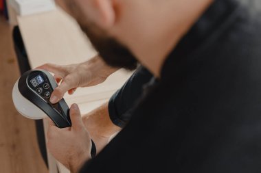 Atletik bir erkek, spor yaptıktan sonra kas ağrısını ve gerilimi azaltmak için parlak bir stüdyoda elektronik bir masaj aleti kullanıyor. Cihaz hedeflenmiş derin doku masajı sağlıyor ve farklı yoğunluk seviyelerine ayarlanabilir..