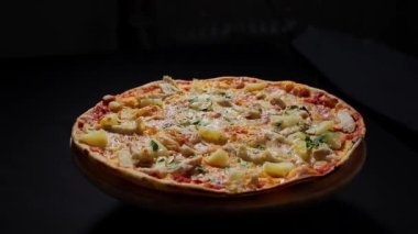 Camera follows the freshly cooked pizza, which is poured over with balsamic sauce. Delicious Italian pizza with chicken and pineapple isolated on a dark background. Traditional Italian food.