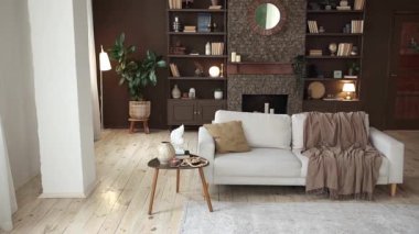 Modern stylish interior with large beige sofa with pillow and plaid on the background of brown wall with brick fireplace, shelving with books and decor and potted plants. Cozy chalet interior. Nobody.
