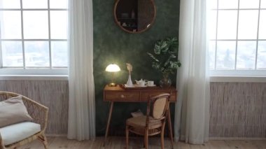 Console table with a table lamp, vase with live plants and a vintage wooden chair in a room with green textured walls with a round mirror, large windows with curtains and a wooden floor. Empty space.