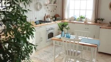 Modern light Scandinavian kitchen with a wooden worktop filled with kitchen accessories. Stylish kitchen with large window, potted plants and wooden dining table with a bouquet of flowers. Nobody.