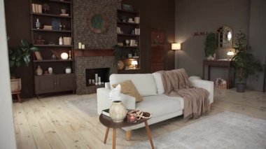 Arka planında kahverengi duvar, tuğla şömine, kitaplar, dekorlar ve saksı bitkileriyle dolu büyük bej koltuklu modern tarz oturma odası. Dağ evinin içi çok rahat. Boş boşluk.