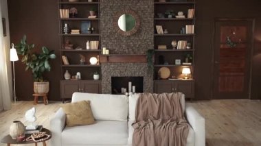 Modern stylish living room with large windows and beige sofa on the background of brown wall with fireplace, shelving with books and decor, and potted plants. Cozy chalet interior. Empty space.