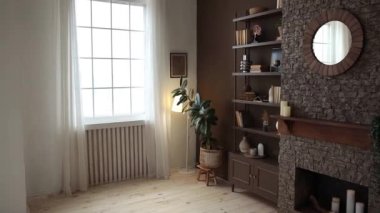 Brown rack with books and decor near the brick fireplace with candles and potted plants in a modern stylish living room with large windows, brown walls and wooden floor. Bookshelf in the home library.