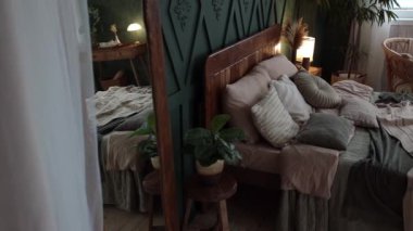 Cozy bedroom interior in boho style with green walls, wooden furniture, console table, vintage chair, craft textiles and carpet, large windows and potted plants. Side view. Nobody.