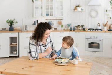 Young beautiful mother feeds her adorable toddler son with healthy vegetables and a chicken cutlet in a bright cozy scandinavian style kitchen. Childs lunch. Mock up, copy space. High quality photo