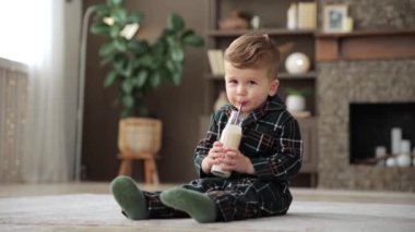 Cute baby drinks milk through an eco-friendly drink straw and sits on the floor. Adorable smiling toddler boy in pajamas is happy to drink a fermented milk drink before his daytime sleep.