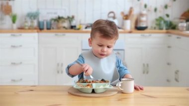 Adorable caucasian toddler boy eats vegetables on his own, pricking them on fork and smiles charmingly. The concept of self-feeding. BLW. Child eats healthy vegetables with meat. Place for text.