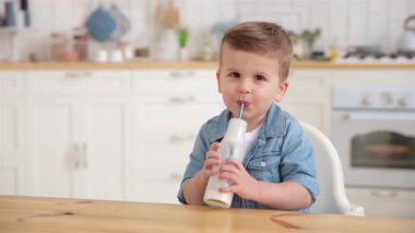 Portrait of cute baby toddler drinks milk or yogurt in the light cozy kitchen. Handsome caucasian boy drinks vitamin rich organic lactose free milk with a straw. Copy space, product mock up. Tasty.