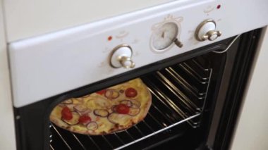 Close up view of young woman takes out a freshly cooked ruddy delicious pizza with mozzarella and tomatoes from the white oven. Homemade pizza cooked in a light spacious Scandinavian style kitchen.