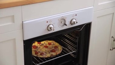 Close up view of young woman takes out a freshly cooked ruddy delicious pizza with mozzarella and tomatoes from the white oven. Homemade pizza cooked in a light spacious Scandinavian style kitchen.