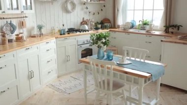 Modern white Scandinavian kitchen with a wooden worktop filled with kitchen accessories. Stylish kitchen interior with large window, potted plants and wooden dining table with white chairs. Nobody.