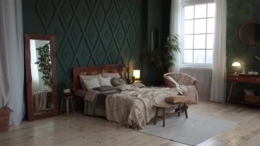 Cozy bedroom interior in boho style with emerald walls, wooden furniture, console table, textiles, large windows and potted plants. Empty space. High quality FullHD footage