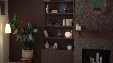 Brown bookshelf with books and decor near the brick fireplace with candles and mirror in a modern stylish interior with large windows, brown walls and potted plants. Wooden rack in the home library.
