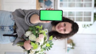 Dikey video. Elinde çiçekleri olan beyaz bir kadın mutfağın bulanık arka planında fotokopi alanı için yeşil ekranlı akıllı telefon tutuyor. Chroma tuşunun modeli. İnternetten alışveriş. Seçici odak.