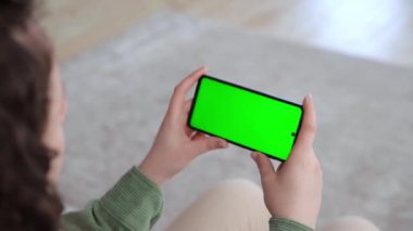 Yeşil ekranlı yatay akıllı telefon kullanan bir kadının arka görüntüsü. Chroma tuşu, yayın hizmetleri uygulamaları için bir model oluşturur. İnternette video izliyorum. Seçici odak.