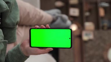 Dikey video. Kanepede uzanmış yeşil ekranlı akıllı telefon kullanan bir kadının bakış açısı. Chroma tuşu reklam uygulamaları ya da yayın hizmetleri için modelleme yapıyor. Seçici odak.