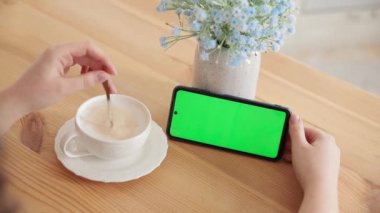 Kadın fotokopi için yeşil ekran kullanıyor. Elinde yatay akıllı telefondan Chroma tuşu modeli var. Kapalı ellerde cep telefonu ve mutfak masasında bir fincan kahveyle. Video izleniyor.