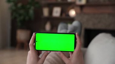 Kadının kanepeye uzanmış yatay akıllı telefon ve yeşil ekran krom anahtar kullandığına dair bakış açısı. Chromakey reklam uygulamaları ya da yayın hizmetleri için modelleme yapıyor. Seçici odak.