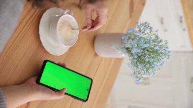 Dikey video. Kadın fotokopi için yeşil ekran kullanıyor. Chroma tuşu akıllı telefondan model alıyor. Kapalı eller cep telefonunu tutar ve mutfak masasında kahve fotograflarını yürütür. İnternetten alışveriş.