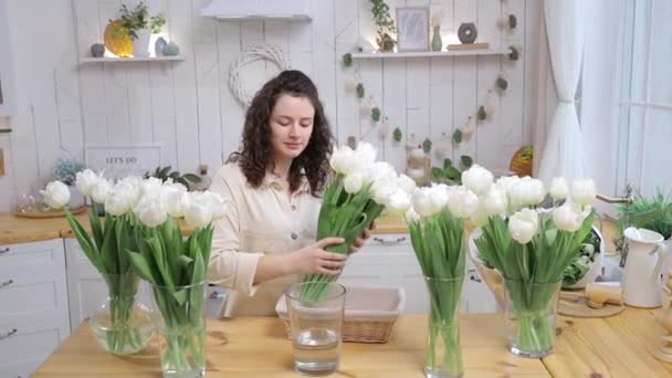 美丽的高加索女人把别致的一束新鲜的白色郁金香放在厨房的花瓶里 手持相机 被初春花朵环绕的女孩 植物区系业务或送花的概念 — 图库视频影像