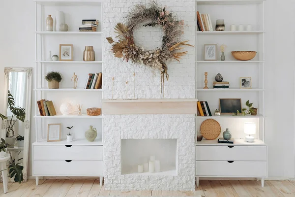 White shelving with books, decor and potted plants near fireplace with wreath of dried flowers in modern stylish living room in scandinavian style. Nobody.