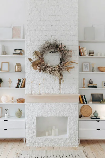 White shelving with books and decor near fireplace with wreath of dried flowers in modern stylish living room in scandinavian style. Nobody.