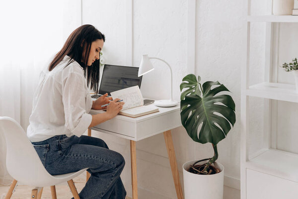 Young brunette freelancer woman sitting at desk using laptop writing notes while studying online, watching webinar, looking at pc screen learning web classes or remote working from home. Side view.