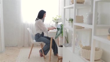 Genç esmer kadın dizüstü bilgisayarda çalışıyor ve arka planda bir buket çiçekle beyaz bir ofis masasında su içiyor. Serbest çalışma kavramı, evde uzak çalışma, çevrimiçi öğrenme.