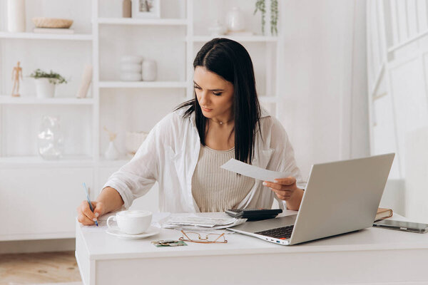 Confident young woman working at desk with laptop, documents, and calculator. Writing notes while reviewing papers. Managing utility payments, taxes, invoices in home office with white shelves.