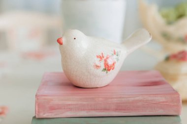 Çiçek süslemeleri ve süslü vazo ile çevrili pastel renkli kitaplara tünemiş narin seramik kuş heykelciği. Yumuşak ışıklar ve yumuşak renkler bir huzur ve cazibe hissi uyandırıyor..