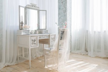 Süslü aynalı şık bir makyaj seti çiçekler ve şamdanla süslenmiş beyaz bir elbise ve üzerine kumaş örtülmüş narin bir sandalyeyle tamamlanmış. Saf perdelerin arasından yumuşak doğal ışık.