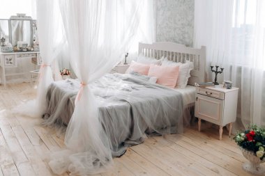 Serene yatak odası beyaz yatak odası ve akan gölgelik, pastel pembe ve gri yastıklar, antika komodin, süslü şamdan ve çiçek buketi. Yumuşak ışık perdelerin arasından süzülür.