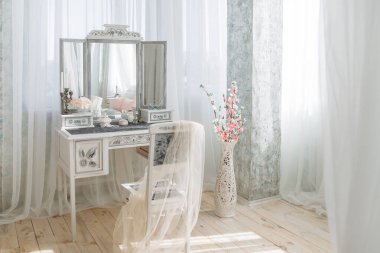 Ayna, beyaz şifonyer, çiçek süslemeleri ve şamdanlı klasik gösteriş. Saf kumaşla örtülmüş narin bir sandalyenin yanında. Yumuşak güneş ışınları katıksız perdelerin arasından huzurlu ve zarif bir ortam yaratır..