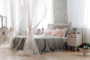 Saf tepe örtüsü ve pastel yastıklı beyaz yataklı zarif bir yatak odası. Kaliteli komodin şamdanı tutarken, yumuşak doğal ışık perdelerin arasından odayı yıkıyor, sükunet ortamını geliştiriyor..