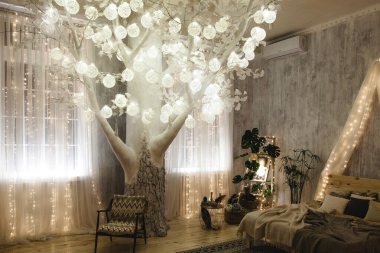 Açık gri duvarlı, rahat bir yatak odasında ışıkları olan dekoratif ağaç, bitkilerle dolu ahşap merdiven rafı, boho tarzı yatak ve hasır sepetler. Oda büyülü ve huzurlu bir atmosfer yaratıyor..