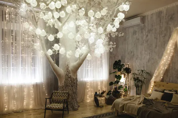Açık gri duvarlı, rahat bir yatak odasında ışıkları olan dekoratif ağaç, bitkilerle dolu ahşap merdiven rafı, boho tarzı yatak ve hasır sepetler. Oda büyülü ve huzurlu bir atmosfer yaratıyor..