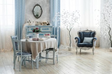 Pastel mavi mobilya, dantel masa örtüsü ve çiçek desenli sofistike iç tasarım. Süslü bir ayna, dekoratif ağaçlar ve yumuşak ışıklandırma odaların cazibesini ve zarafetini arttırır..