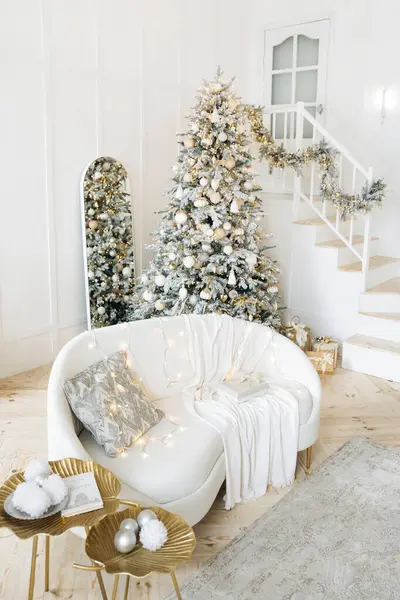 Merdivenleri olan modern bir oturma odası, gümüş ve altınla süslenmiş bir Noel ağacı, yastıklı ve battaniyeli beyaz bir kanepe, ağacın altında hediyeler. Oda bayram sıcaklığı ve neşe saçıyor..