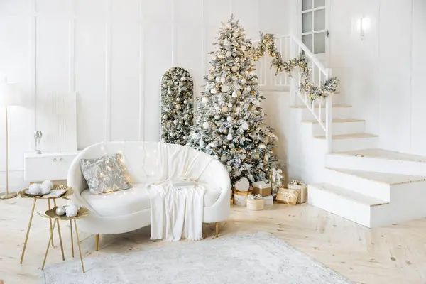 Gümüş ve altınla süslenmiş bir Noel ağacı olan modern oturma odası yastıklı ve battaniyeli beyaz kanepe, ağacın altında hediyeler. Oda bayram sıcaklığı ve neşe saçıyor..