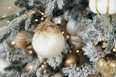 Buzlu bir Noel ağacının üzerindeki beyaz ve altın bir süslemenin yakın plan çekimi. Ilık ışıklar, şenlikli bir parıltı ekliyor. Karmaşık tasarım ve parlak detaylar lüks bir tatil havası yaratıyor..