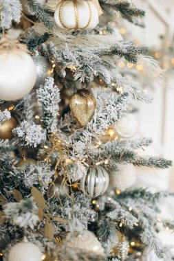 Beyaz ve altın süslemelerle süslenmiş buzlu Noel ağacı dallarının yakın çekimi. Parlak ışıklar, bayram dekorunun şenlikli ve lüks atmosferini artırır..