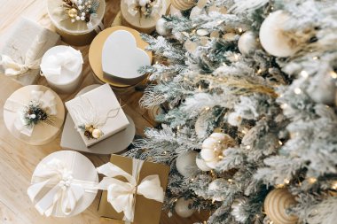 Altın ve beyazla süslenmiş, fiyonklarla süslenmiş ve güzel süslenmiş bir ağacın altına yerleştirilmiş zarif Noel hediyeleri. Ilık ışıklandırma rahat bir tatil ortamı yaratır..