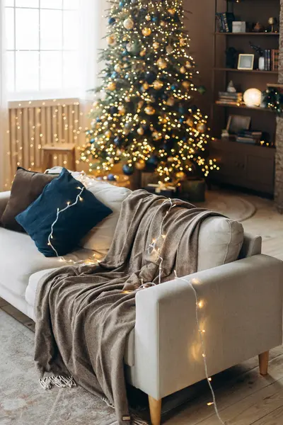 Yastıklı bir kanepe ve arka planda bulanık bir ekoseyle büyük pencereli ve kahverengi duvarlı rahat bir oturma odasının iç tarafında çelenk ışıkları ile süslenmiş bir Noel ağacı. Sıcak Noel Arifesi.