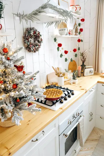 Ahşap mutfak atölyesi ve üzerinde Noel pastası olan gaz ocağı. Noel ağacı olan İskandinav mutfağı çelenk ışıkları ve kırmızı oyuncaklar ve duvarında bir koni çelengi.