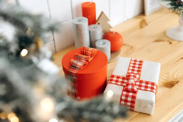 Noel ağacının altında tahta bir masanın üstünde kırmızı kareli kurdeleli yeni yıl hediyeleri. Yakın çekim, seçici odaklanma, bulanık bir Noel ağacından çelenklerin ışıklarından Bokeh 'i görmek..