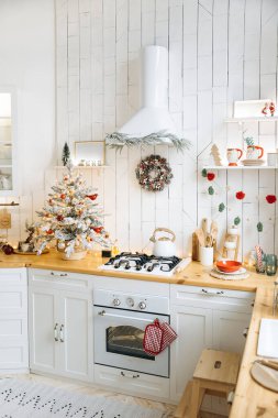 Beyaz dolaplar ve ahşap tezgahlarla süslü mutfak. Kırmızı süslemeler, çelenkler, çelenkler ve sobanın üzerinde beyaz bir çaydanlıkla süslenmiş küçük bir Noel ağacı..