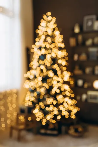 Yumuşak odaklı altın ışıklar, rahat bir oturma odasında Bokeh olan bir Noel ağacına. Tatil için dekore edilmiş arka plan rafları neşeli havayı arttırır. Sığ DOF.