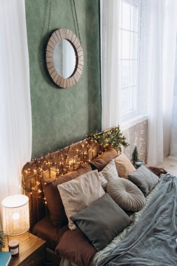 Ahşap yatak başlığı, peri ışıkları ve yastıklar, beyaz perdeli büyük pencereler tarafından yerleştirilmiş, yuvarlak aynalı yeşil duvara karşı.